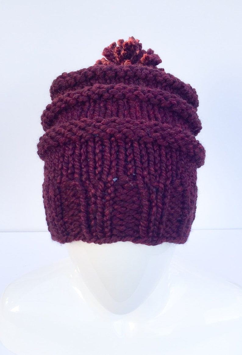 May include: A burgundy knitted beanie with a pom pom on top. The beanie has a cable knit pattern.