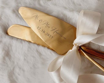 May include: A gold-toned cake knife and server set featuring a serrated edge and a cream-colored satin ribbon tied at the handles. The server is engraved with the text "Mr & Mrs Harrison" and the date "12.10.2027" in a cursive script font.