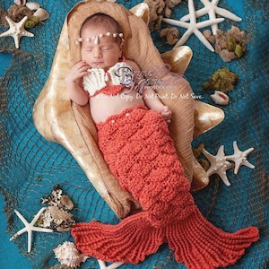 May include: A newborn baby wearing a crocheted mermaid tail costume, lying in a seashell prop. The baby is wearing a pearl headband and holding a seashell. The background is a blue and white fishing net with starfish and seashells.