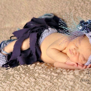 May include: A newborn baby wearing a dark blue fabric skirt and a matching headband with a decorative flower and veil. The baby is lying on a textured, light brown surface.