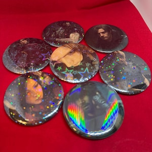 May include: Six circular pin-back buttons featuring various character portraits. The buttons have a holographic finish with star-shaped and rainbow-coloured light reflections across the surfaces, displaying different figures in dark, moody tones.
