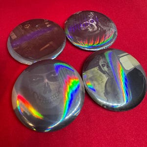 May include: Four circular pin-back buttons featuring holographic finishes that reflect rainbow light. Each button displays a distinct graphic of a skull-masked figure in dark tones. The designs include close-up portraits and tactical gear imagery.