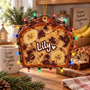 May include: A banana bread slice ornament featuring chocolate chips, two banana slices, and the name Lily with a heart printed in white. The ornament is wrapped in a string of small, multi-colored LED Christmas lights and hangs from a gold cord.