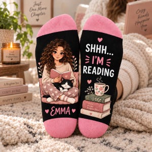 May include: A pair of black socks with pink cuffs and toes. One sock features an illustration of a person with curly hair reading a book with a cat, and the name Emma. The other sock displays the text SHHH... I'M READING, a mug, and books.