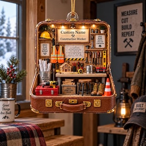 Könnte beinhalten: Eine Miniatur-Bauwerkstatt in einem braunen Koffer-Ornament. Es enthält ein Schild mit individuellem Namen, eine Warnweste, einen Holzhausrahmen, Werkzeuge, einen Hocker und Arbeitsstiefel. Der Innenraum ist mit kleinen Lichtern beleuchtet.
