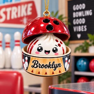 May include: A hanging ornament shaped like a red glittery bowling ball that opens to reveal a white dumpling character with a smiling face. The base features the text Brooklyn in black script, flanked by two white bowling pins on a tan background.