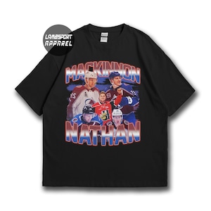 May include: A black short-sleeved t-shirt featuring a graphic print of hockey player Nathan MacKinnon in various jerseys. The text MACKINNON and NATHAN appears in a red, white, and blue gradient font. A circular logo reads LAMBSPORT APPAREL.