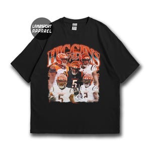 May include: A black short-sleeve t-shirt featuring a graphic print of football players in orange and white uniforms. The text "HIGGINS" appears in large orange letters above the players, and "LAMBSPORT APPAREL" is printed in a black circle.