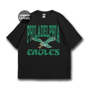 Può includere: Una t-shirt nera a maniche corte con una grafica verde di un'aquila che tiene un pallone da football. Il testo PHILADELPHIA è arcuato sopra l'aquila e la parola EAGLES è stampata sotto in verde. Un logo circolare riporta LAMBSPORT APPAREL.