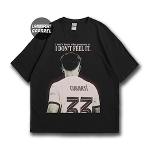 May include: A black short-sleeved t-shirt featuring a graphic of a football player from the back wearing a light-coloured jersey with the name CUBARSÍ and the number 33. Above the player, the text reads: i don't know what pressure is. I DON'T FEEL IT.