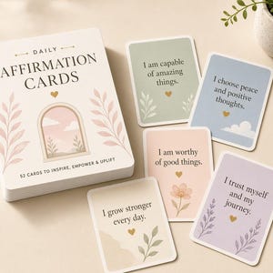 Può includere: Un mazzo di carte di affermazioni quotidiane con una scatola che presenta un'illustrazione ad arco e il testo 52 CARDS TO INSPIRE, EMPOWER & UPLIFT. Quattro carte singole mostrano frasi come I am capable of amazing things, accompagnate da icone a forma di cuore dorato e motivi botanici.