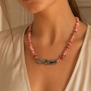Puede incluir: Un collar de cuentas compuesto por una hilera de pequeñas piedras irregulares de color rosa y melocotón. El centro del collar está acentuado con tres piedras lisas, rectangulares y de color gris verdoso iridiscente, separadas por pequeños espaciadores metálicos.