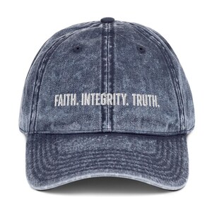 May include: A navy blue acid-wash baseball cap featuring the embroidered white text FAITH. INTEGRITY. TRUTH. centred on the front panel. The cap has a six-panel construction with a curved brim and a button at the top centre of the crown.