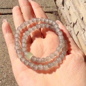May include: A stretch bracelet composed of smooth, translucent grey spherical beads. The bracelet features a single, slightly larger, pale green square-shaped bead integrated into the circular strand of grey beads, creating a subtle contrast.