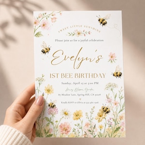 May include: A white birthday invitation card featuring watercolour illustrations of bees and wildflowers. The text reads: Sweet Little Sunshine, Evelyn's 1st Bee Birthday, Sunday, April 13 at 3:00 PM, Honey Blossom Garden, 87 Meadow Lane, Spring Hill, CA 90218.