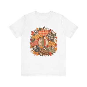 Puede incluir: Una camiseta blanca de manga corta con un gráfico central de un grupo de calabazas con varios patrones, incluyendo cuadros, estampado de leopardo, cachemir y lunares, decorado con pequeños fantasmas blancos, hojas y flores.