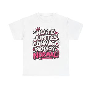 Puede incluir: Una camiseta blanca de manga corta con un estampado gráfico que incluye el texto NO TE JUNTES CONMIGO NO SOY NORMAL en letras blancas gruesas con bordes rosas y negros, decorado con pequeños corazones rosas, una corona y un icono de cara sonriente.