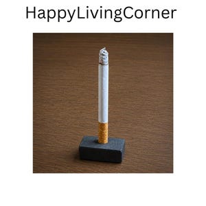以下が含まれることがあります： 白いフィルターとベージュの紙の本体を持つ火のついたタバコが、小さな長方形のマットブラックの台座に垂直に立っています。オブジェクトの上にはHappyLivingCornerというテキストがあります。タバコの先端には灰色の灰がついています。