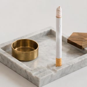 Puede incluir: Un cenicero cuadrado de mármol blanco con un inserto circular de latón, un cigarrillo apoyado en una muesca tallada y una pieza decorativa de madera geométrica unida a la esquina de la bandeja, mostrando vetas de piedra natural.