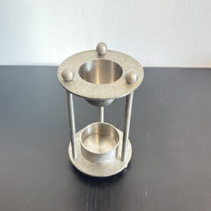 May include: A silver-toned metal candle holder featuring a circular top plate with three small spherical accents, supported by three vertical rods. The structure includes a central upper bowl and a lower circular base with a small cup for a candle.