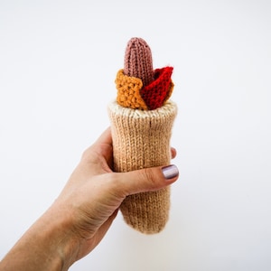 Könnte beinhalten: Eine Hand hält einen gestrickten Hotdog mit Senf und Ketchup. Der Hotdog ist aus Garn und hat eine hellbraune Farbe. Der Senf und der Ketchup sind ebenfalls aus Garn und sind gelb und rot.