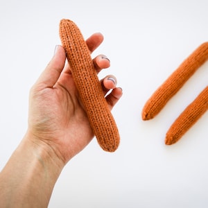 Könnte beinhalten: Drei gestrickte orangefarbene Karotten. Eine Karotte wird in einer Hand gehalten. Die Karotten sind etwa 25 cm lang.