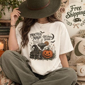 Könnte beinhalten: Ein cremefarbenes Kurzarm-T-Shirt mit einem schwarzen Grafikdruck und dem Text Practical Magic Halloween Season Midnight Margaritas. Das Design zeigt einen Hexenhut, einen Bücherstapel, ein Margaritaglas, einen Kürbis und Fledermäuse.