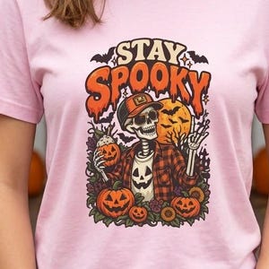 May include: A light pink t-shirt featuring a graphic print of a skeleton wearing a plaid shirt and a baseball cap. The skeleton holds a small pumpkin, surrounded by larger pumpkins, sunflowers, and bats. The text STAY SPOOKY is printed above.
