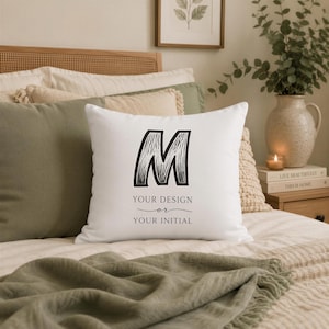 May include: A square white throw pillow featuring a large black letter M in a sketched font style. Below the letter, the text reads YOUR DESIGN or YOUR INITIAL in a clean, black serif typeface, centered on the front of the pillow cover.