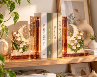 May include: A pair of amber-colored acrylic bookends featuring a crescent moon, stars, and lily of the valley floral designs. The bookends hold four books titled The Kinfolk Home, At Home, The Art of Home, and Live Beautifully.