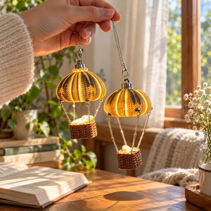 May include: Two hanging ornaments shaped like hot air balloons, each featuring a yellow and dark gray sea urchin shell as the balloon canopy, connected by silver chains to a small brown woven basket containing light-colored stones.