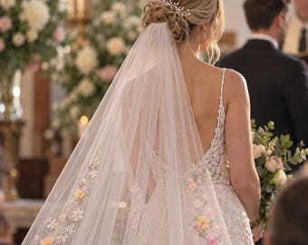 Può includere: Un lungo velo da sposa bianco e trasparente con delicati ricami floreali colorati lungo i bordi, abbinato a un abito in pizzo bianco senza maniche con scollo sulla schiena e un accessorio per capelli argentato con perline e cristalli.
