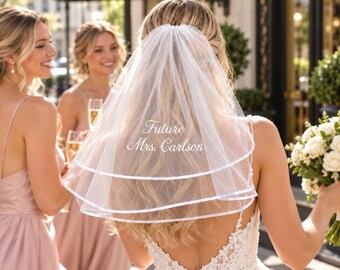 Può includere: Un velo da sposa bianco in tulle a due livelli con un bordo in nastro di raso lungo i bordi. Il velo è ricamato con il testo bianco Future Mrs. Carlson centrato sul tessuto. Il materiale trasparente ricade su lunghi capelli biondi.