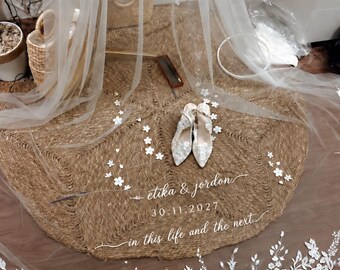 Può includere: Un velo da sposa bianco trasparente con intricati ricami in pizzo floreale bianco lungo i bordi e applicazioni floreali sparse. Il velo riporta il testo "etika & jordon", "30.11.2027" e "in this life and the next" in corsivo bianco.