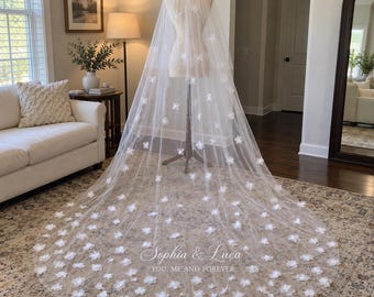 Può includere: Un lungo velo da sposa bianco e trasparente drappeggiato su un manichino, caratterizzato da applicazioni floreali bianche tridimensionali sparse su tutto il tessuto. Il testo "Sophia & Luca YOU, ME AND FOREVER" è stampato in bianco sul bordo.