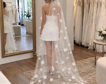 Può includere: Un abito corto bianco senza spalline abbinato a un lungo velo in tulle trasparente decorato con applicazioni floreali bianche tridimensionali sparse. Il velo è fissato con una fascia floreale coordinata, creando un look armonioso.