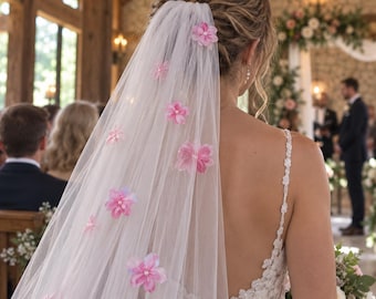 Può includere: Un velo da sposa bianco in tulle trasparente, decorato con fiori in tessuto rosa sparsi con piccole perle bianche al centro. Il velo è fissato a un'acconciatura raccolta e ricade sulla schiena sopra un abito in pizzo bianco.