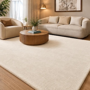 May include: A large, rectangular area rug in a solid cream color with a soft, textured pile. The rug is positioned on a light brown wooden floor, extending across the foreground of a living room space with neutral-toned furniture.