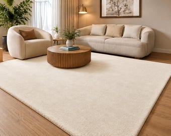 Könnte beinhalten: Ein großer, rechteckiger Teppich in einfarbigem Creme mit weichem, strukturiertem Flor. Der Teppich liegt auf einem hellbraunen Holzboden und erstreckt sich über den Vordergrund eines Wohnbereichs mit Möbeln in neutralen Tönen.