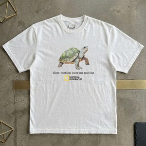 Puede incluir: Una camiseta blanca de manga corta con un gráfico de una tortuga verde y marrón. Debajo de la tortuga, el texto dice "slow motion over no motion" seguido del logotipo amarillo y negro de National Geographic centrado en el pecho.