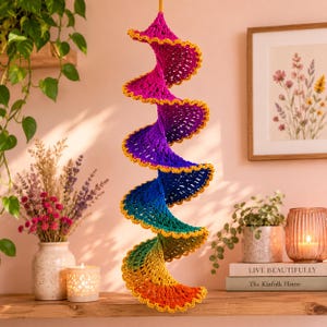 May include: A hanging spiral decoration made of crocheted yarn in a vibrant rainbow gradient, transitioning from magenta at the top to orange at the bottom. The edges of the spiral are finished with a decorative yellow scalloped trim.