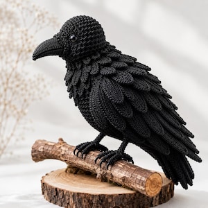 May include: A black crocheted raven figurine featuring detailed layered wing feathers, a textured beak, and a glossy black eye. The bird is perched on a small wooden branch attached to a circular wooden slice base, showcasing intricate needlework.