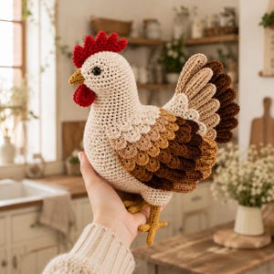 May include: A crochet chicken toy featuring a cream-colored body, a bright red comb and wattle, and layered wing feathers in shades of light brown, medium brown, and dark brown. The chicken has a small yellow beak and yellow crocheted feet.