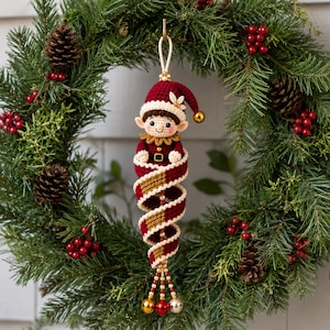 May include: A crocheted elf ornament featuring a deep red hat with a gold bell, a cream-coloured face, and a dark red and gold spiral body. The ornament is suspended by a cream cord and finished with small gold, red, and silver dangling bells.