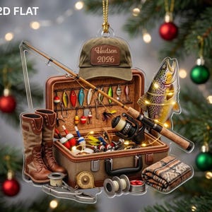 May include: A flat, decorative ornament featuring a fishing theme with a brown baseball cap labeled Hudson 2026, a tackle box filled with colorful lures, a fishing rod, brown boots, a plaid blanket, and a trout, all arranged in a festive display.