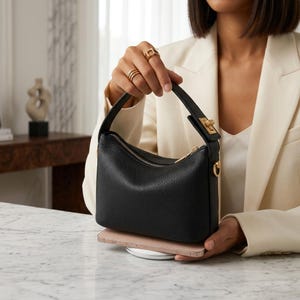 May include: A black pebbled leather handbag with a single top handle, a gold-toned metal clasp closure on the strap, and a top zipper. The bag is positioned on top of a rose gold smartphone, showcasing its compact size and structured design.