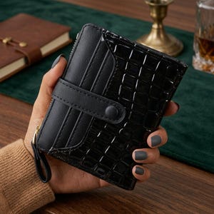 Peut inclure: Un portefeuille noir à deux volets présentant une texture gaufrée façon crocodile brillante sur le corps principal et une finition mate lisse sur la section porte-cartes. Il comprend une patte de fermeture à bouton-pression et une tirette dorée.
