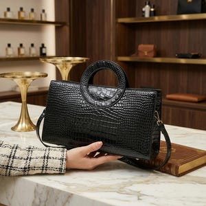 May include: A black handbag with a glossy crocodile skin texture, featuring a structured rectangular body and a prominent circular top handle. The bag includes a thin, detachable shoulder strap attached to the sides with small gold-toned metal hardware.