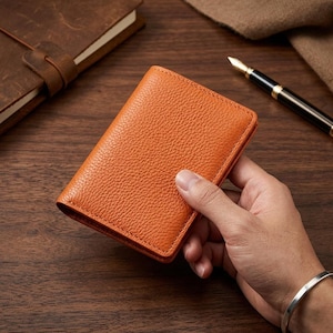 May include: A vibrant orange leather card holder featuring a distinct pebbled texture. The rectangular accessory has clean, stitched edges and a folded design, showcasing a bright, saturated color that contrasts with the surrounding dark wood surface.