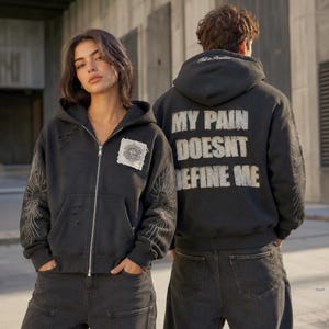 Pode incluir: Um casaco preto com capuz e fecho, detalhes de tecido desgastado, um emblema quadrado no peito e desenhos gráficos de raios de sol nas mangas. As costas exibem o texto "MY PAIN DOESNT DEFINE ME" em letras grandes com aspeto envelhecido.